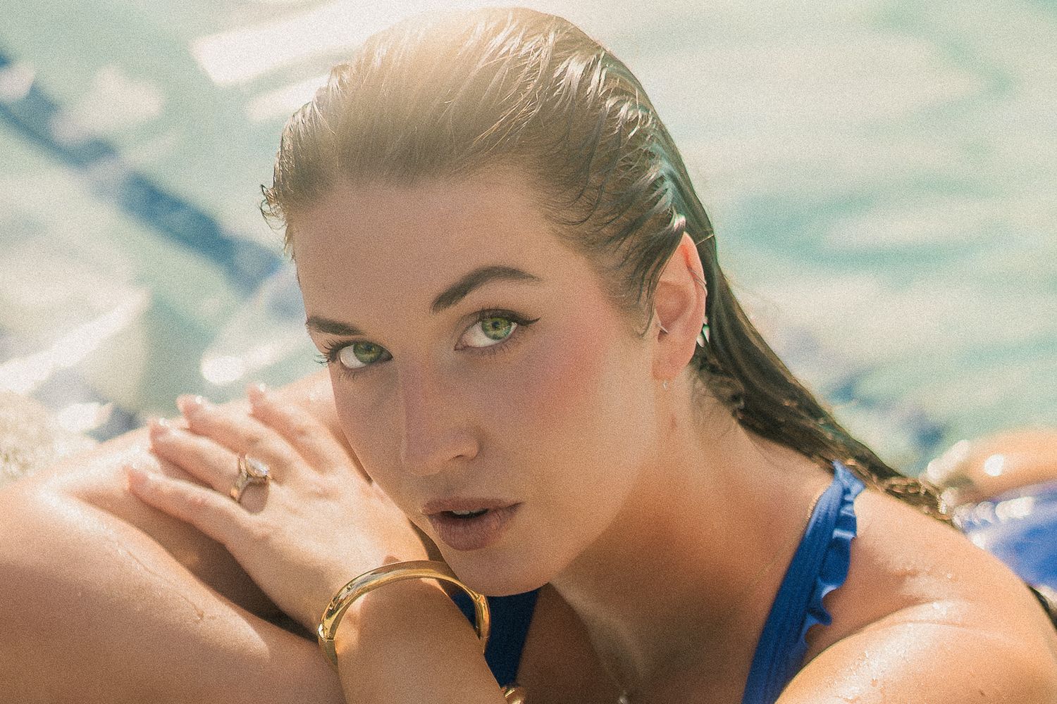 A woman with green eyes poses poolside in a blue bikini with golden jewelry in sunlight.