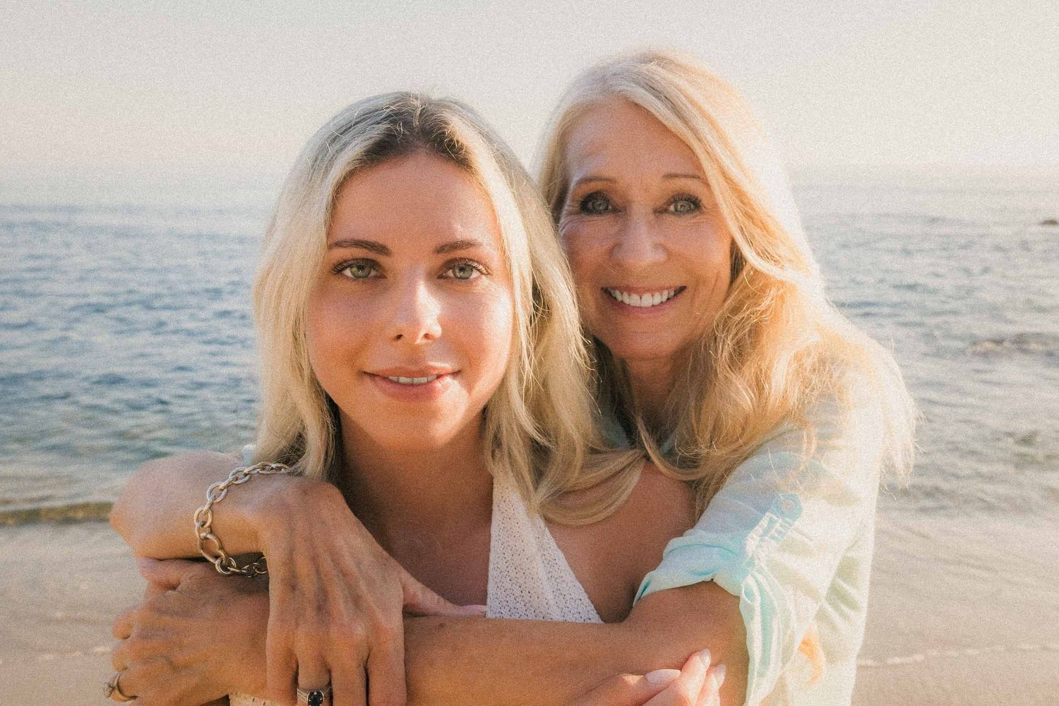 Two blonde women smile warmly together on a sunny beach with ocean views behind them.