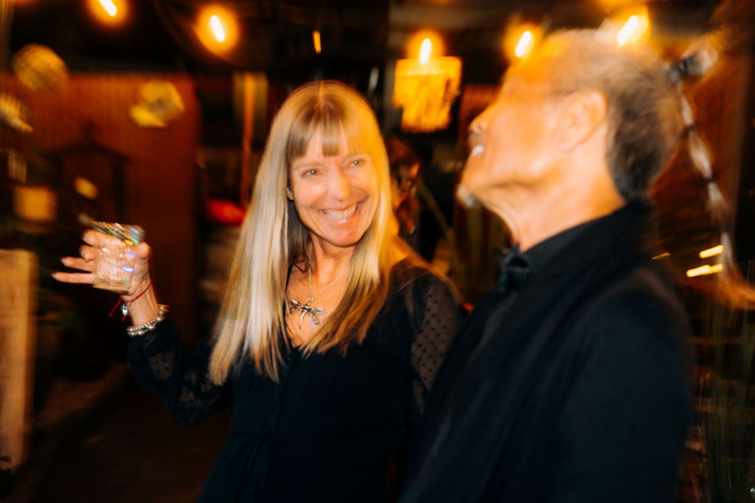 Smiling blonde woman holding a cocktail drink at a warmly lit social gathering.