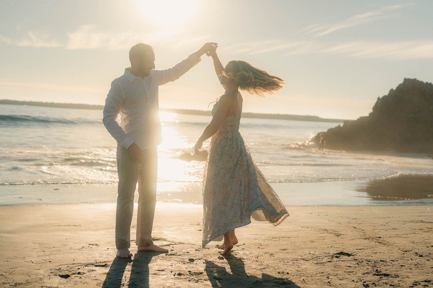 Couple dancing on golden sunset beach, man spinning woman in flowing dress by the ocean.