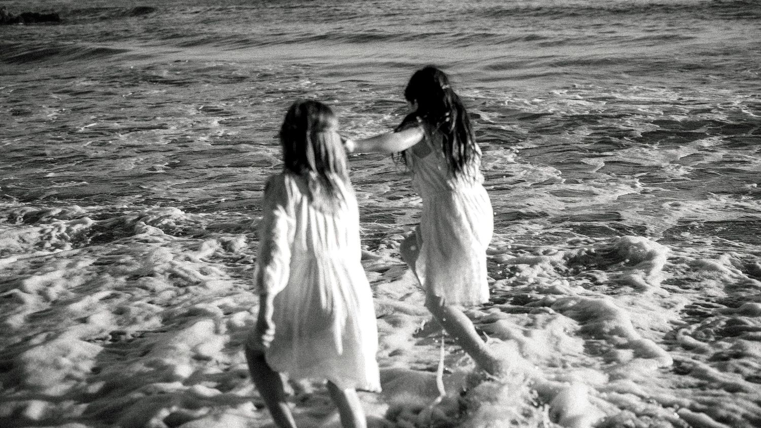Black and white photo of two girls in white dresses wading in ocean waves.