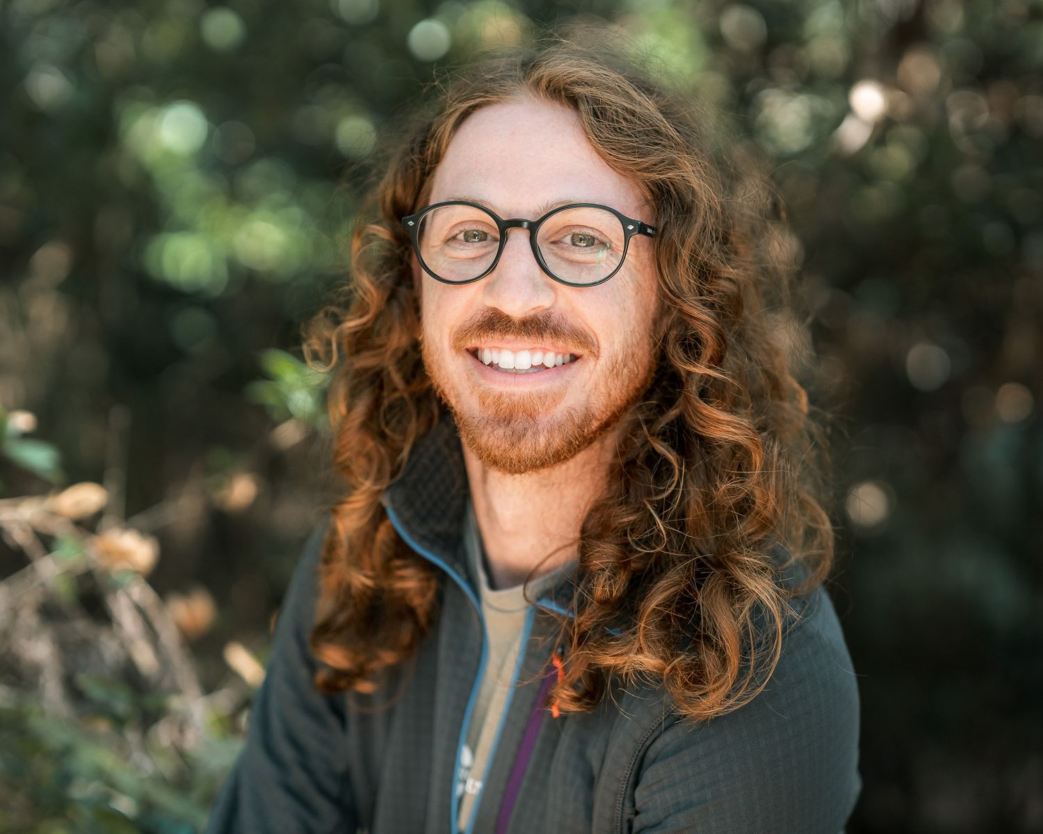 A young man with curly hair and round glasses smiles warmly in a natural outdoor setting.