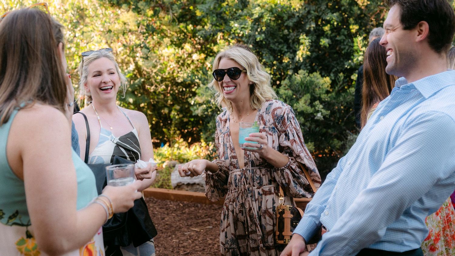 Group of women laugh and enjoy drinks together at a sunny outdoor garden party event.