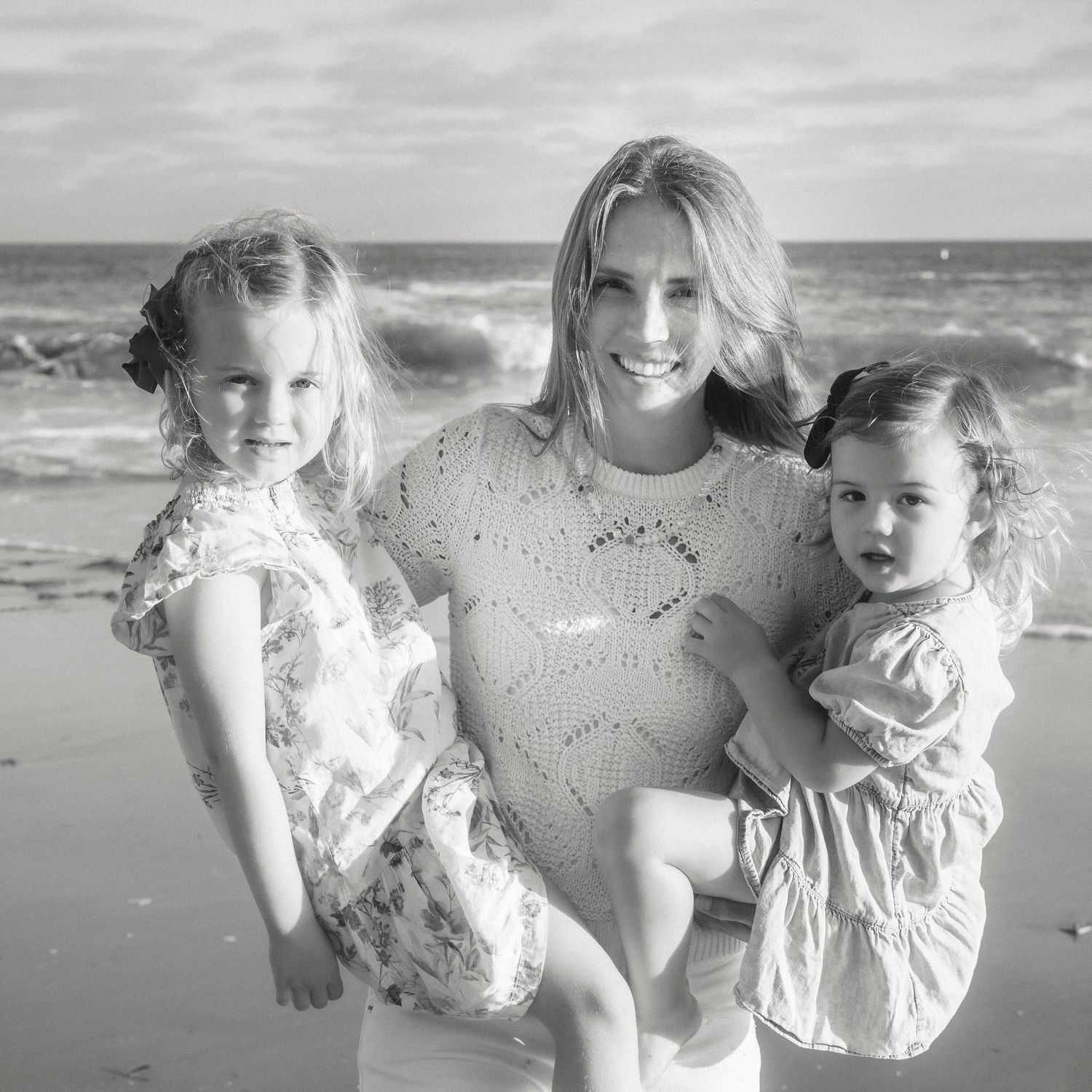 Black and white photo of a woman holding two young girls on a beach with ocean waves behind them.