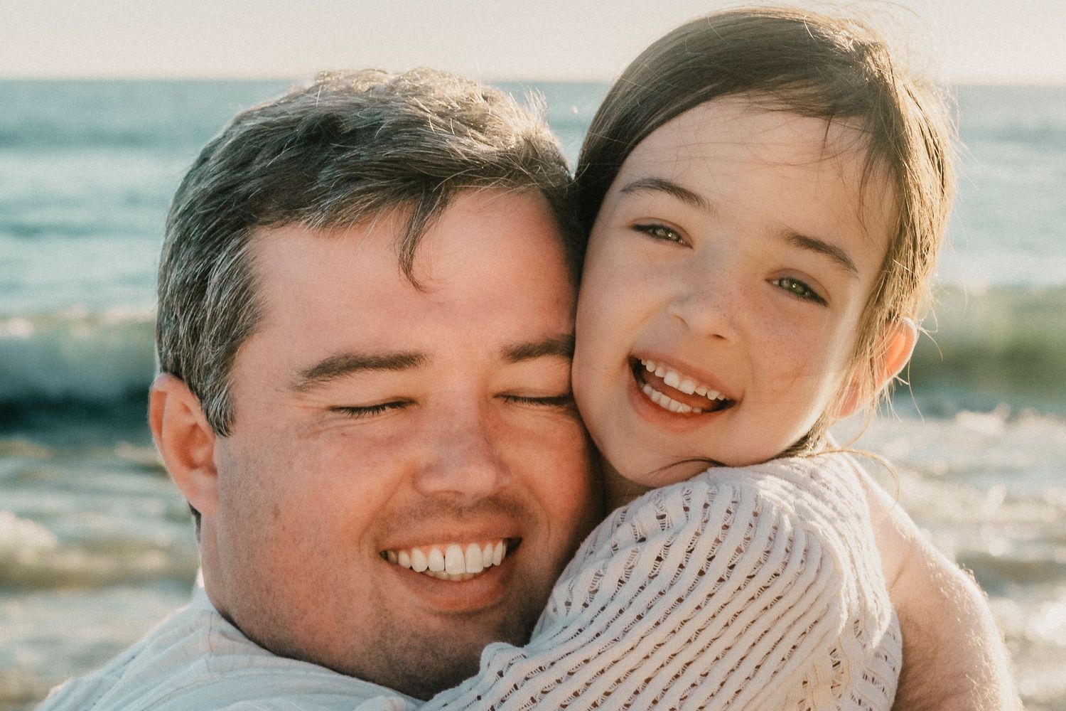 A smiling father and daughter share a joyful hug on a sunny beach.