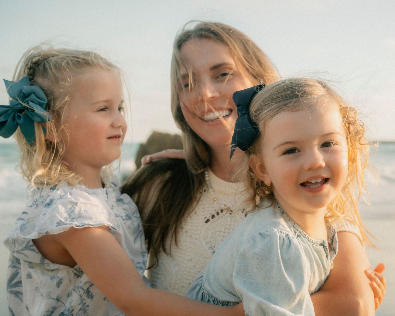 Blonde mother smiling with two young daughters wearing hair bows at sunny beach.
