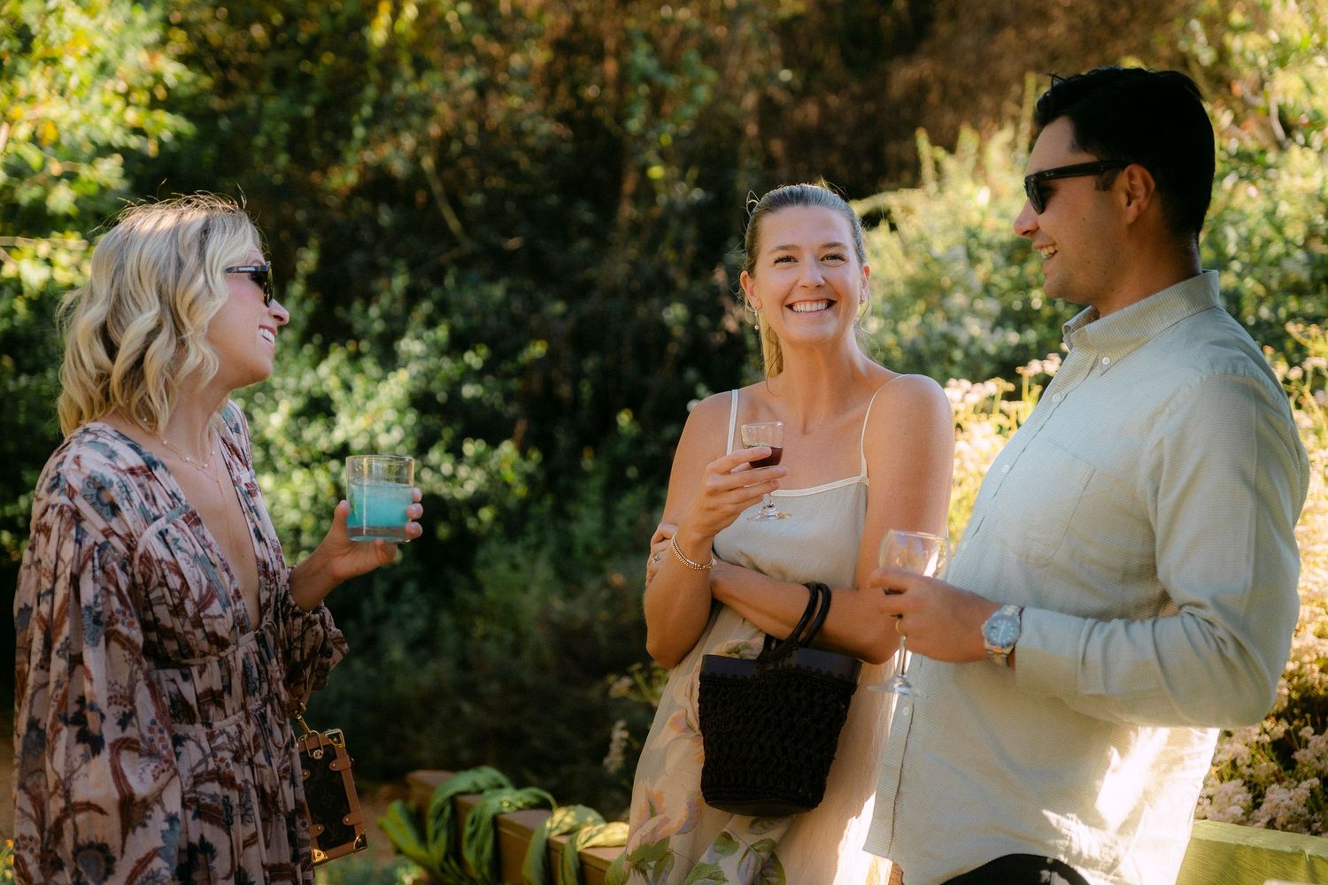 Three friends laughing and socializing outdoors while holding drinks in a lush garden.