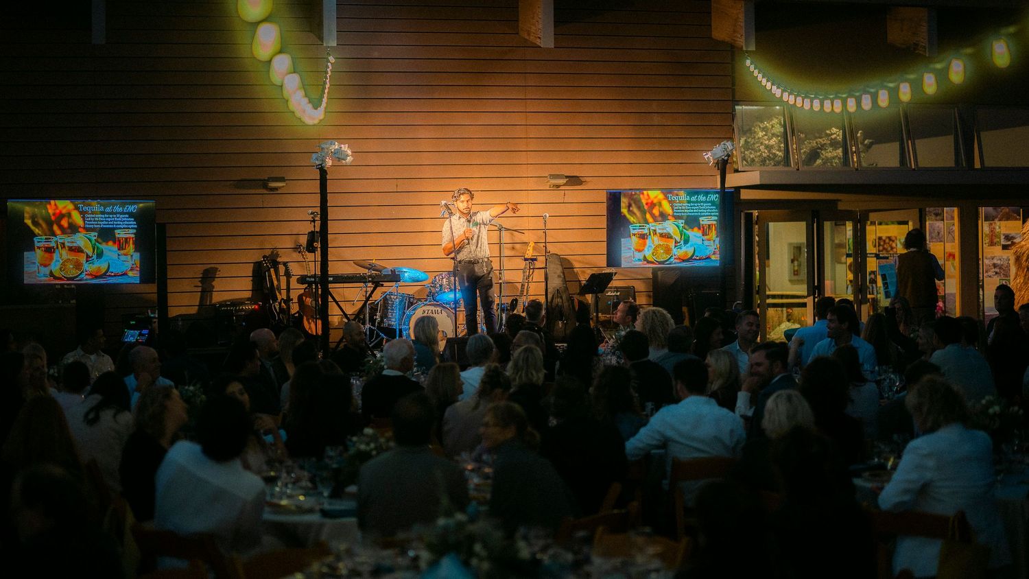 Performer on stage with colorful screens entertaining a seated crowd at an outdoor night event.