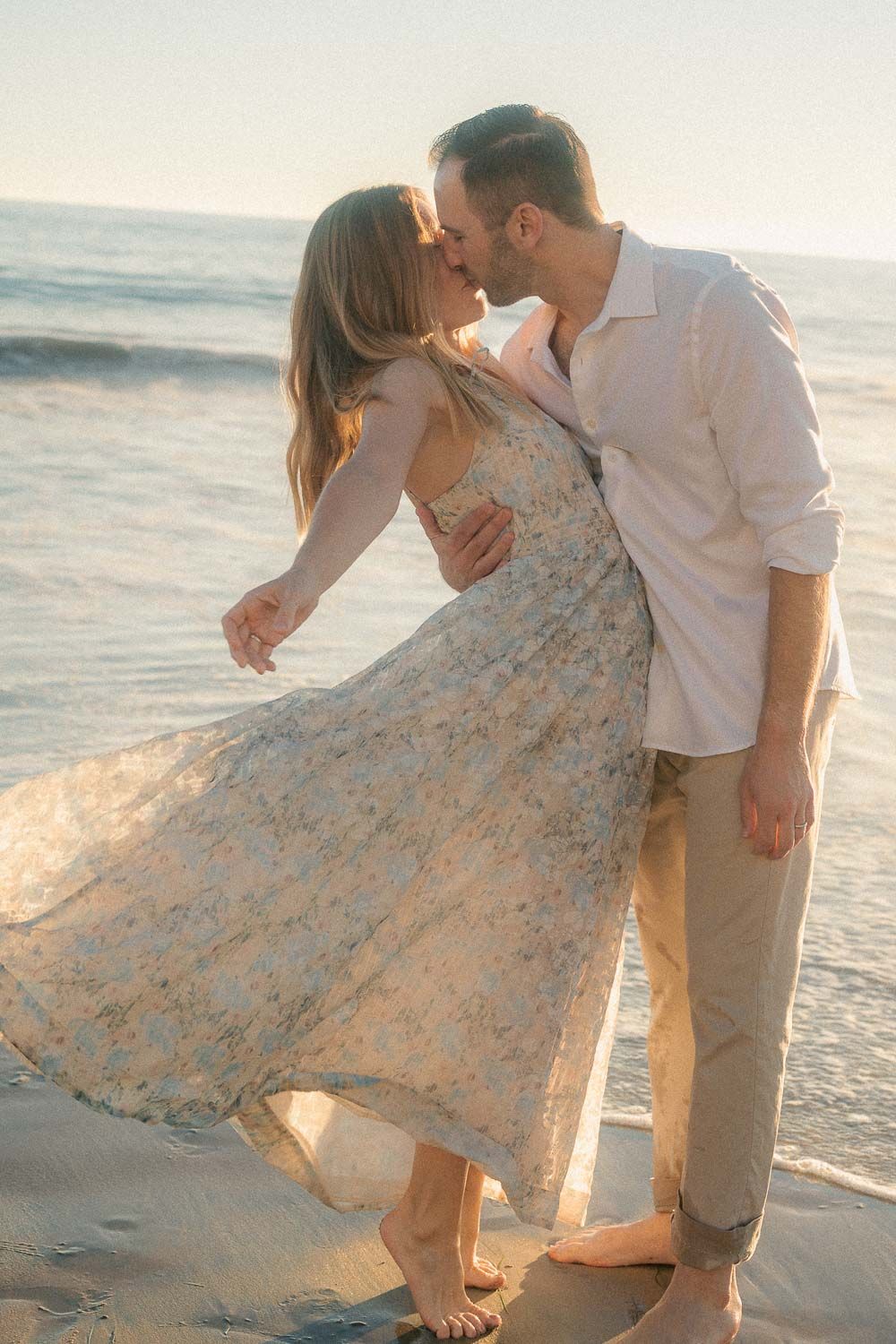 Couple sharing a romantic kiss on the beach at sunset, woman in flowing floral dress with ocean waves behind them.