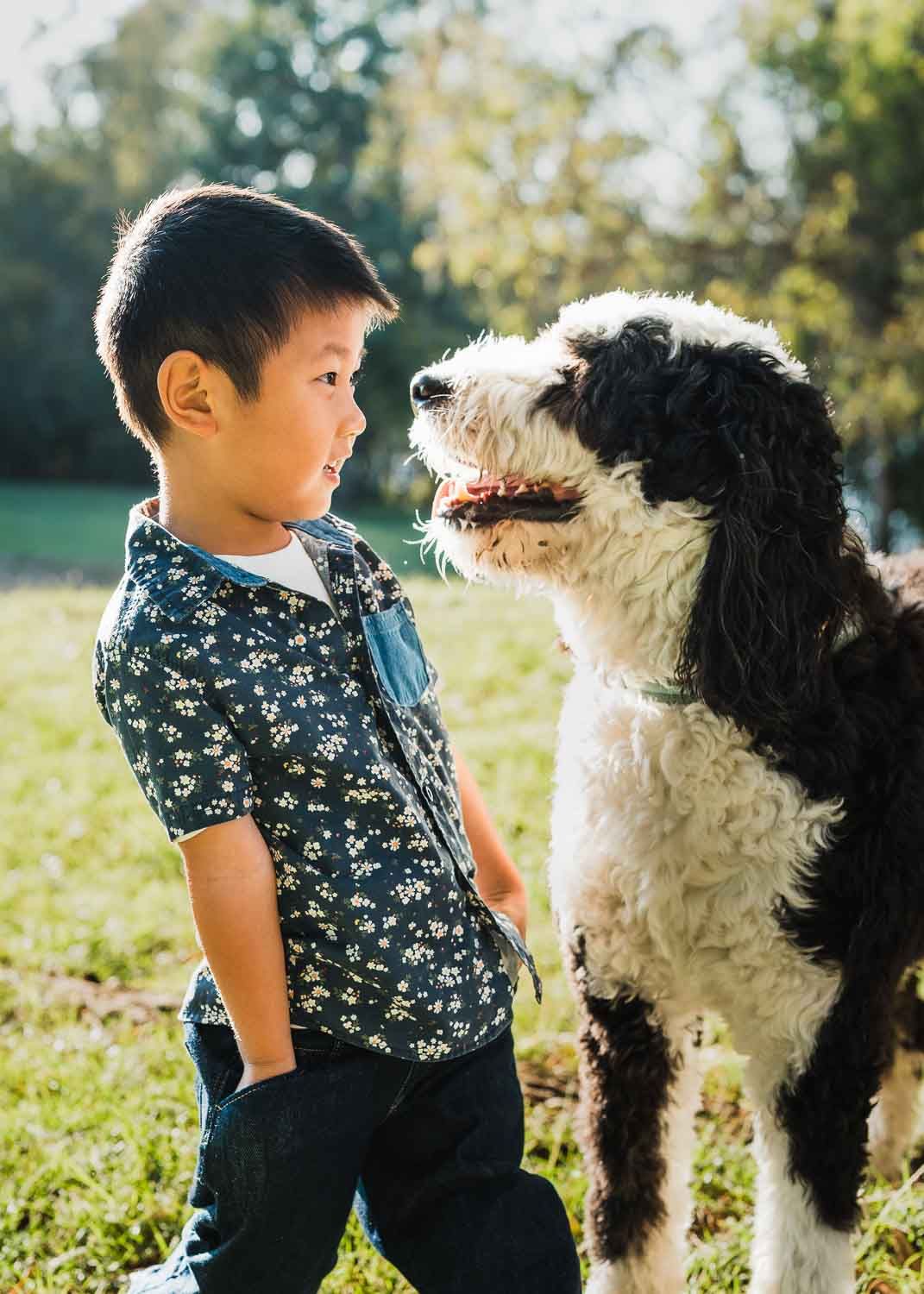 Smiling young Asian boy in floral shirt standing with a black and white dog outdoors.