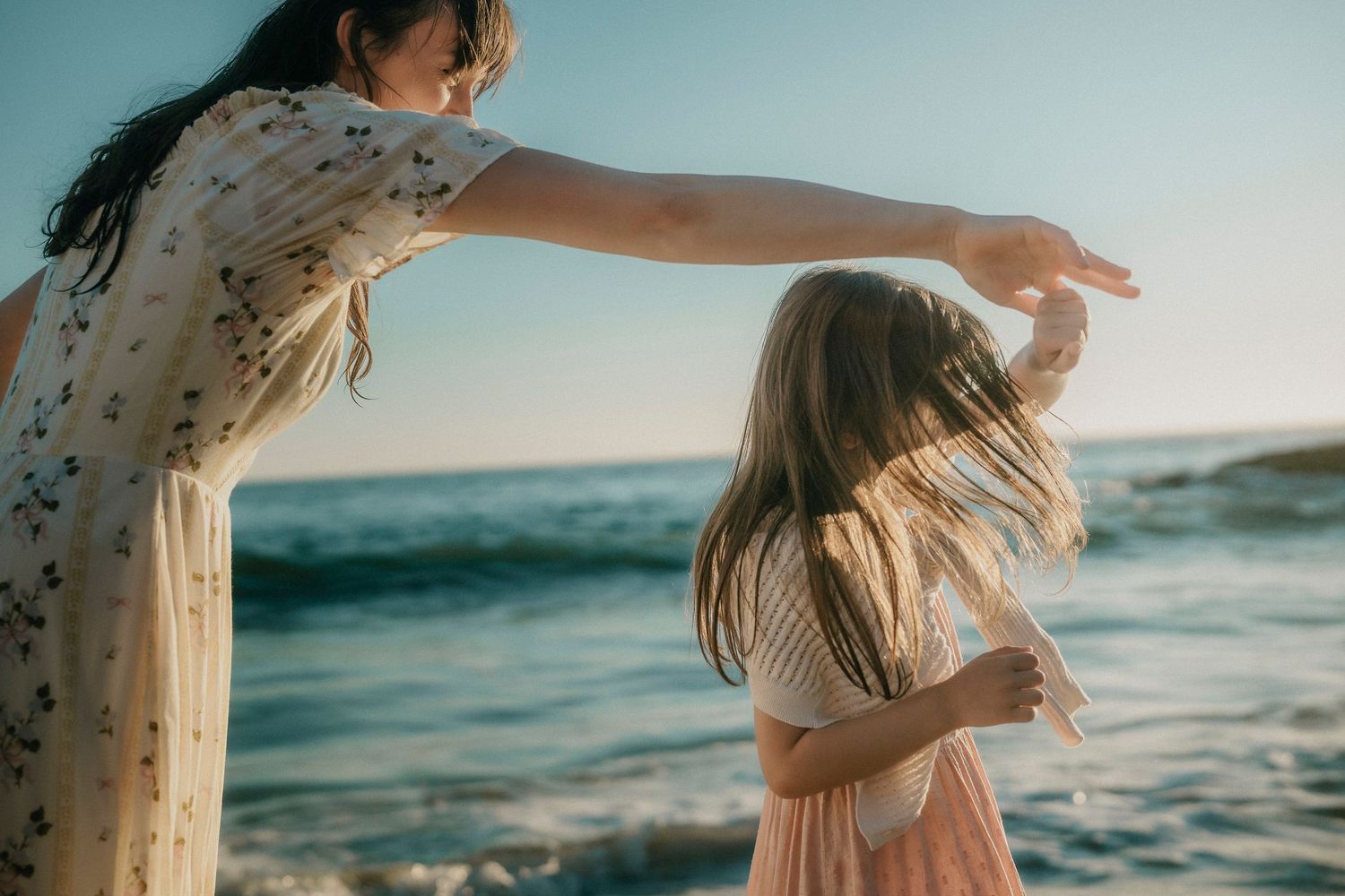 Mother spinning her daughter by the ocean shore during a golden hour beach photoshoot.
