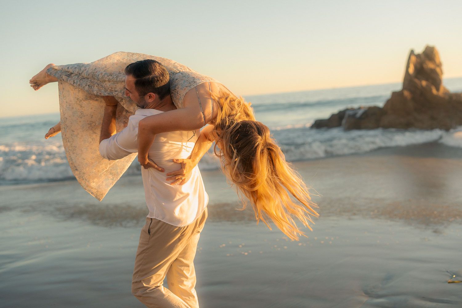 Man lifting woman joyfully on beach at golden sunset with rocky coastline and ocean in background.