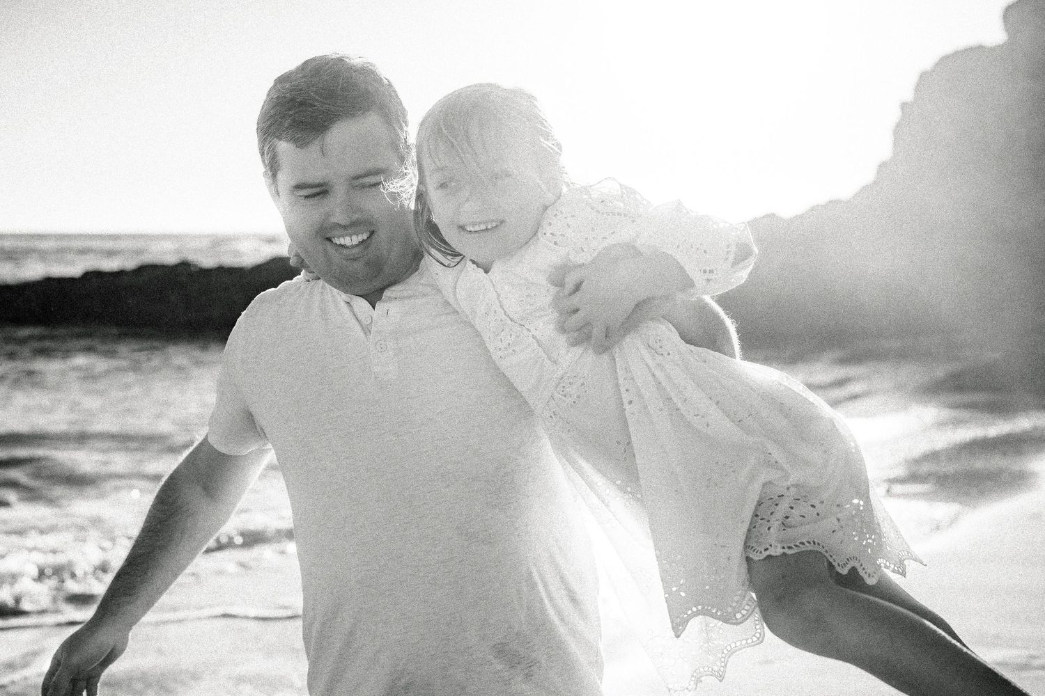 Black and white photo of a laughing father carrying his daughter on a beach.