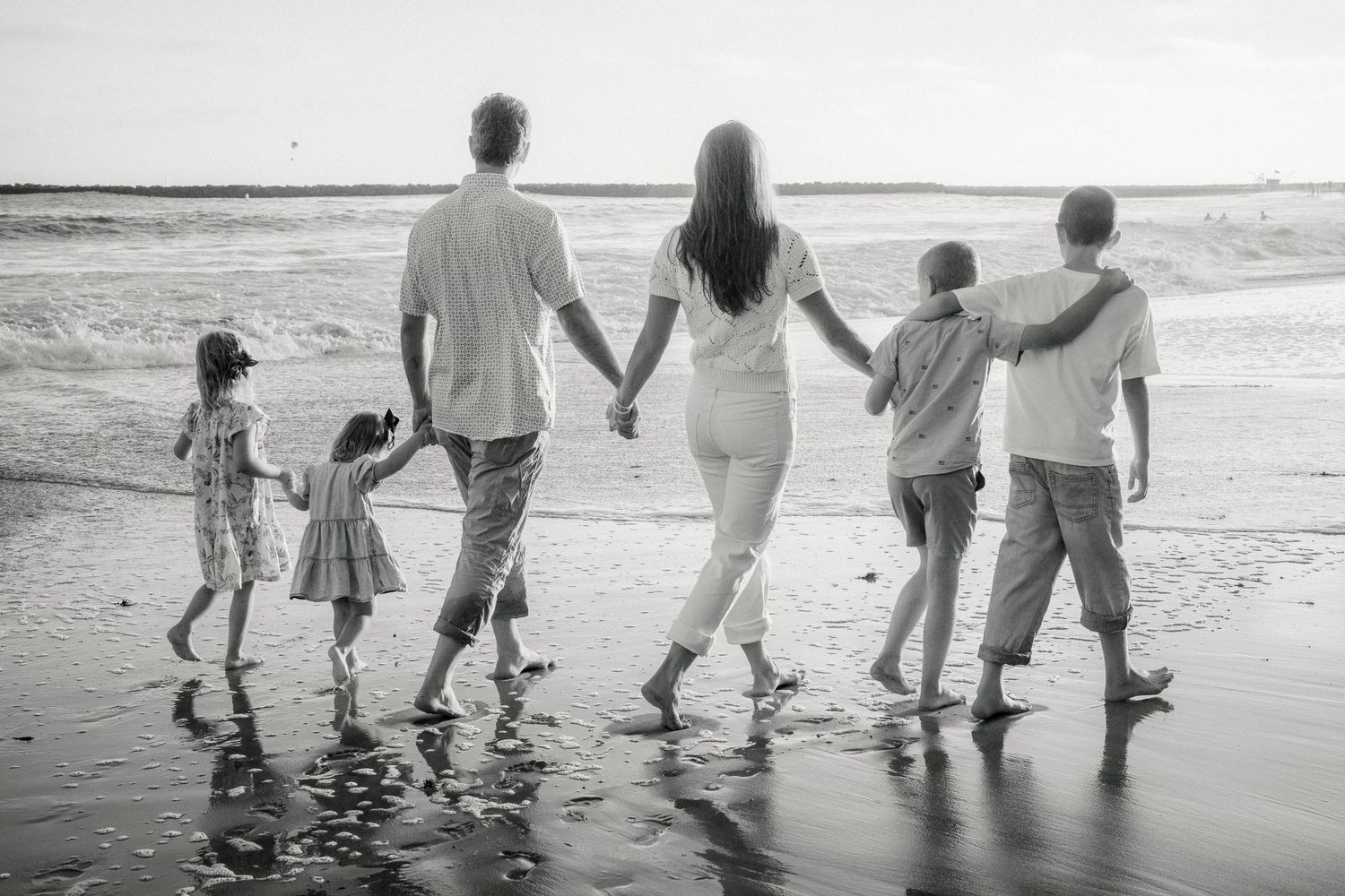 Black and white photo of a family of six walking hand-in-hand along a beach shoreline.