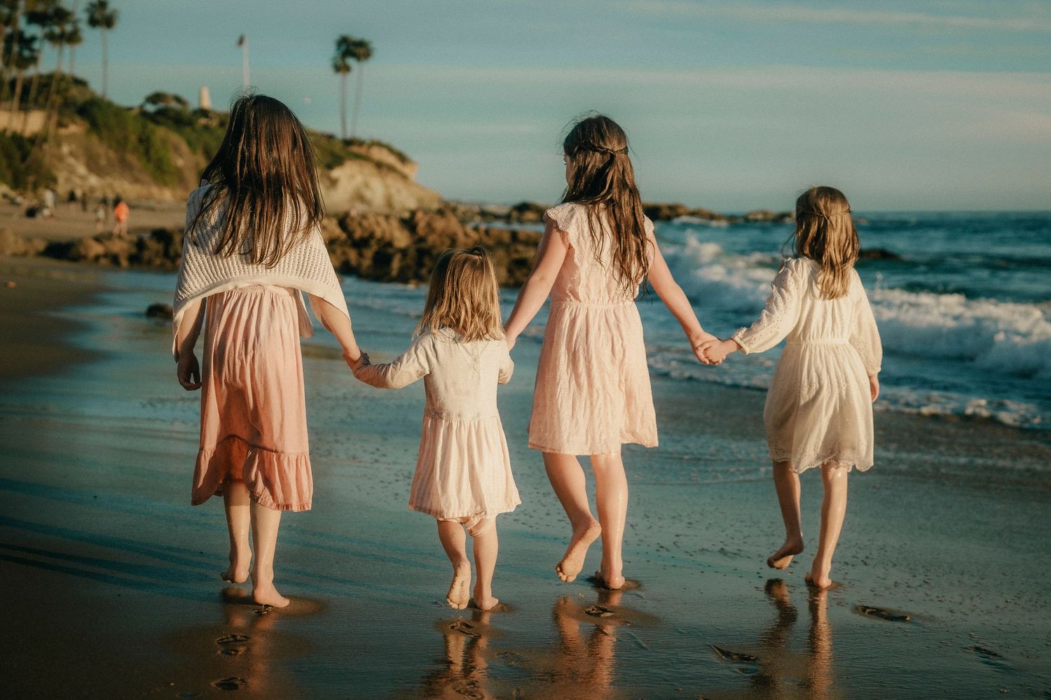 Four girls in pastel dresses holding hands walking along a sandy beach shoreline.