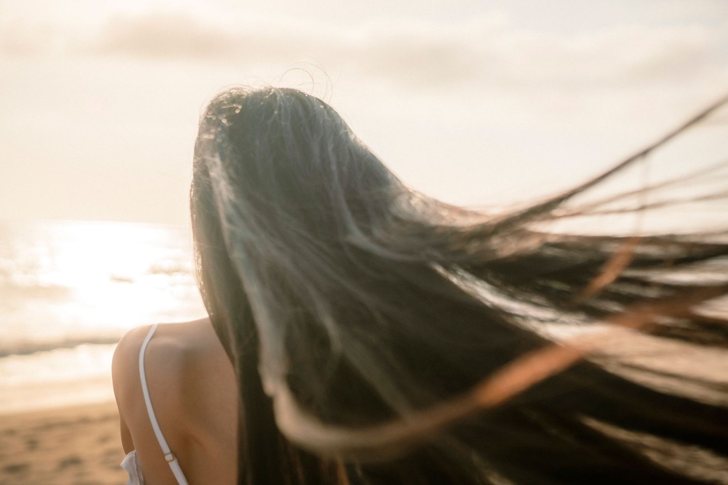 A woman with long dark hair blowing in the wind stands on a beach during golden hour sunset.