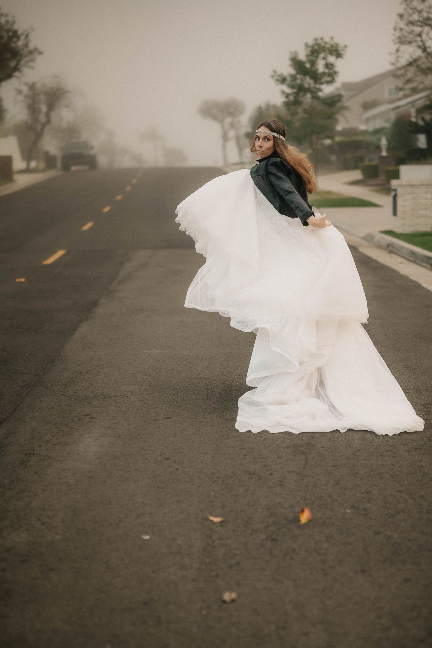 Bride in white wedding gown and black leather jacket twirling on a foggy suburban street.