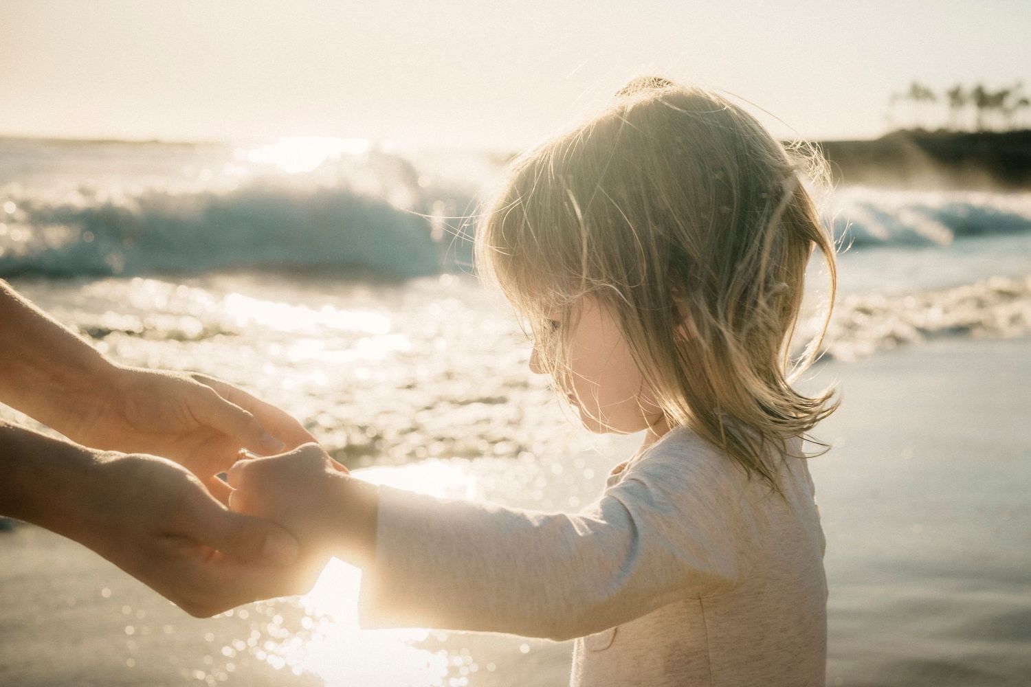 A young child holds an adult's hands on a sunlit beach with waves in the background.