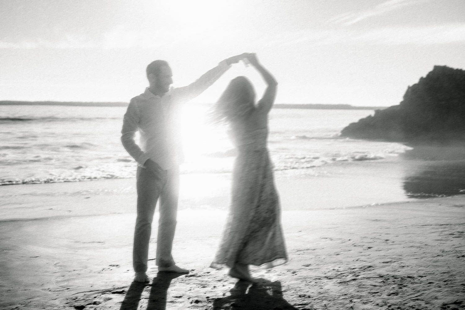 Black and white silhouette of couple dancing on sunlit beach with ocean waves behind them.