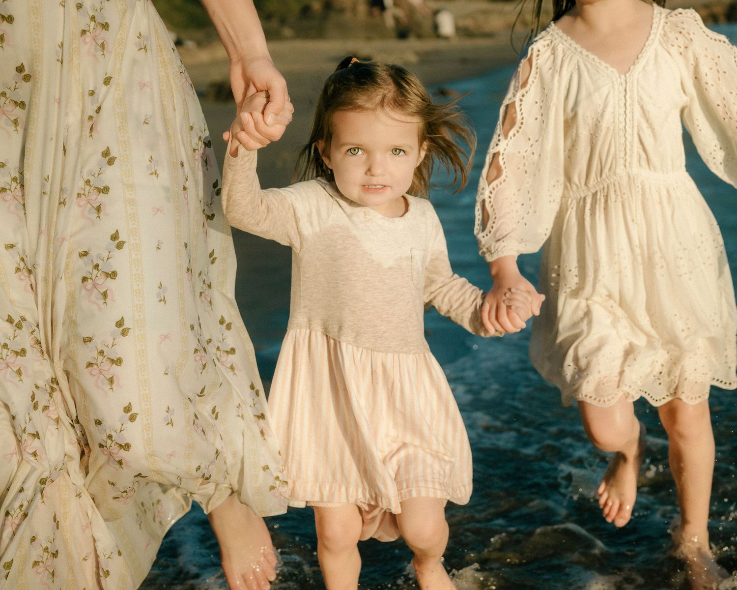 Young toddler girl in cream dress holding hands with adults while wading in ocean.