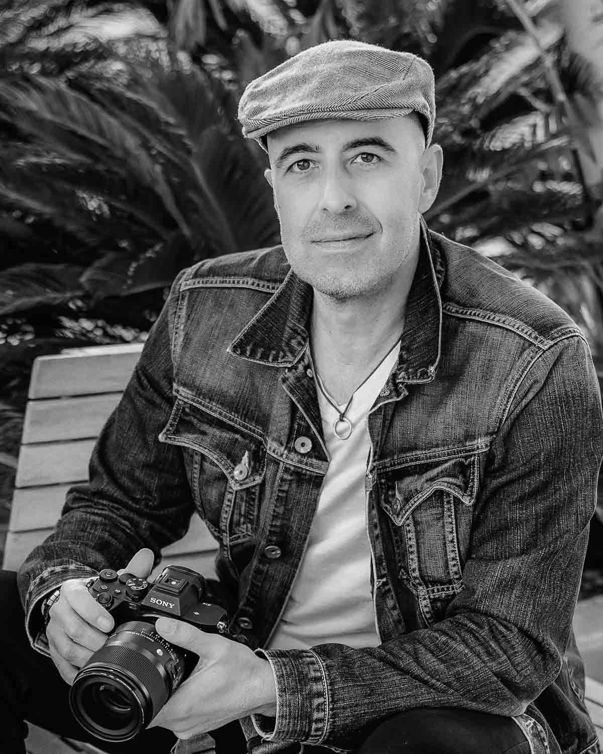 Black and white portrait of smiling male photographer sitting with Sony camera near palm trees.