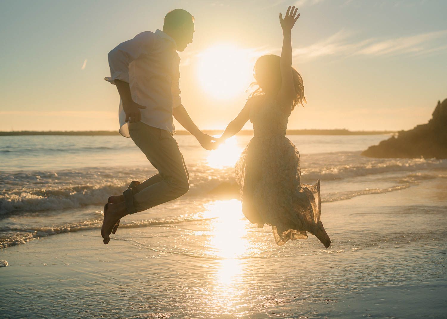Joyful couple jumping together on beach at sunset, silhouetted against glowing golden sun over ocean.