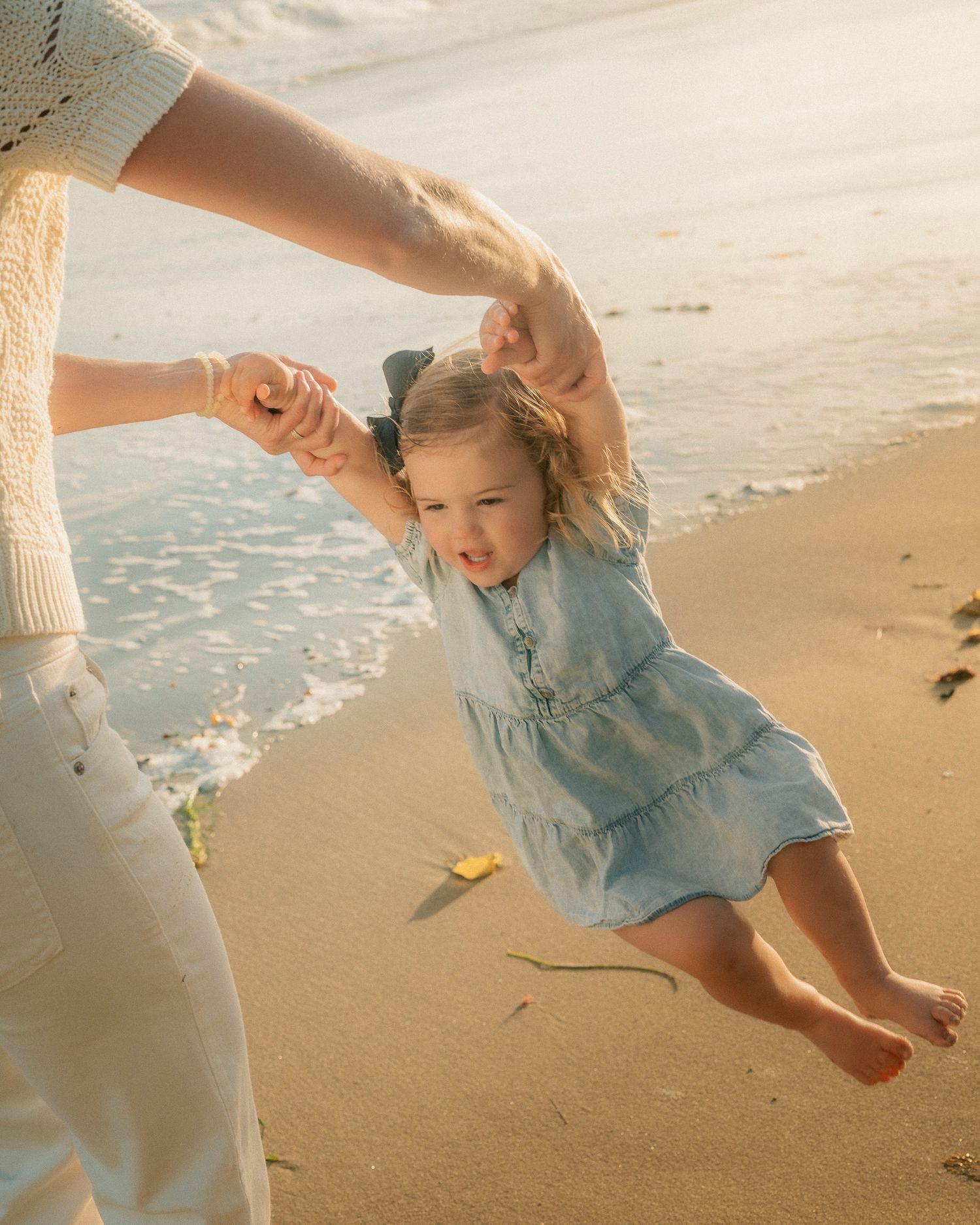 Adult swinging happy toddler girl by hands on sandy beach near ocean waves.