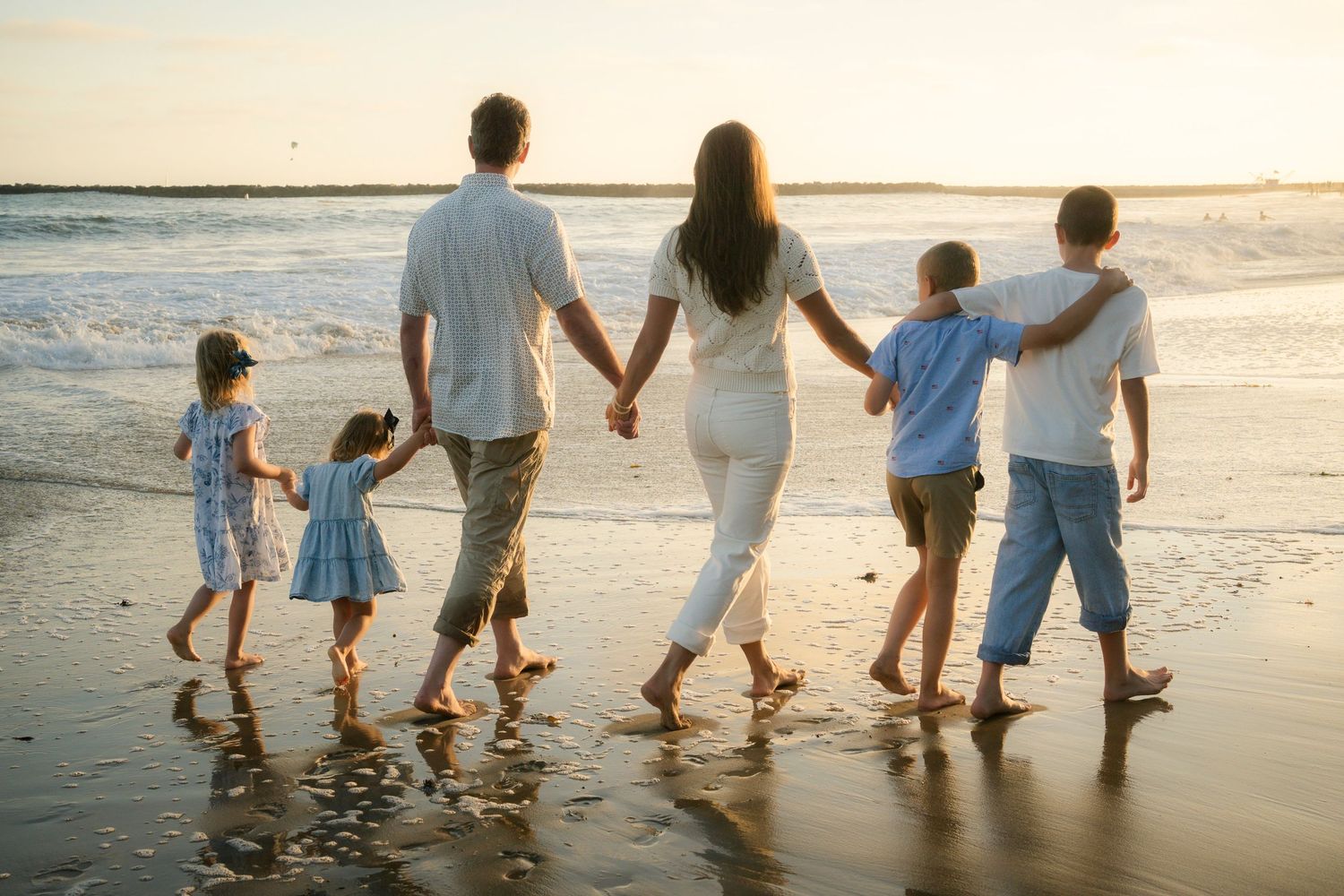 Family of six walking hand-in-hand along a sunlit beach shoreline viewed from behind.