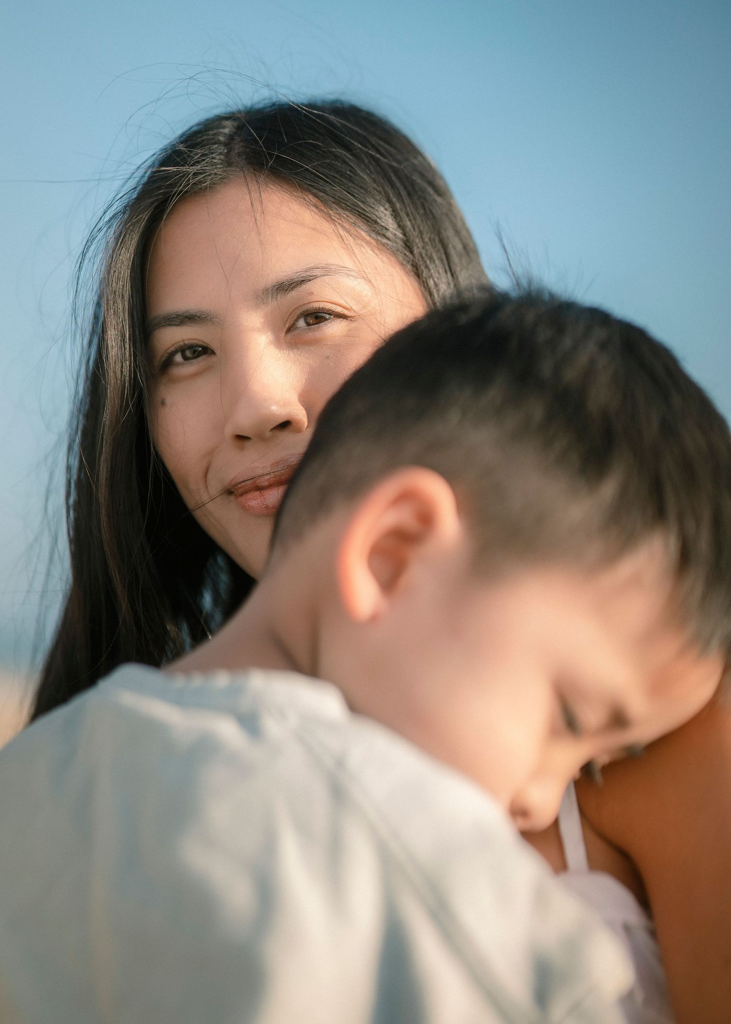 Woman with long dark hair gazing at camera while holding a young boy at the beach.