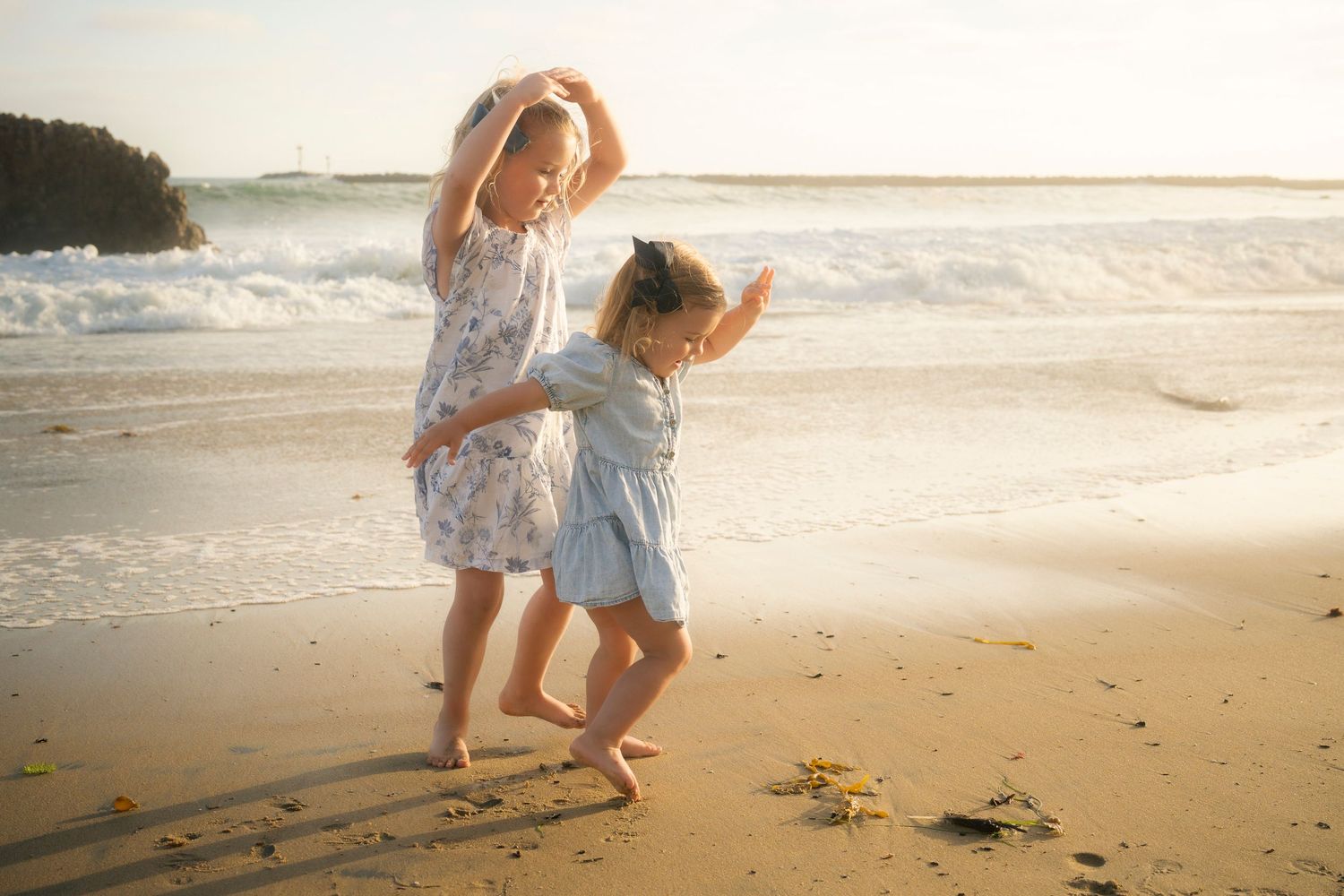 Two young girls in dresses dancing playfully on a sunny beach.