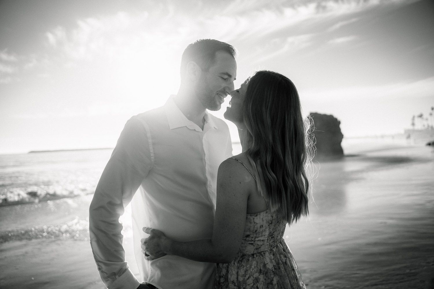 Black and white romantic couple embracing on beach with dramatic sunlight and ocean waves.