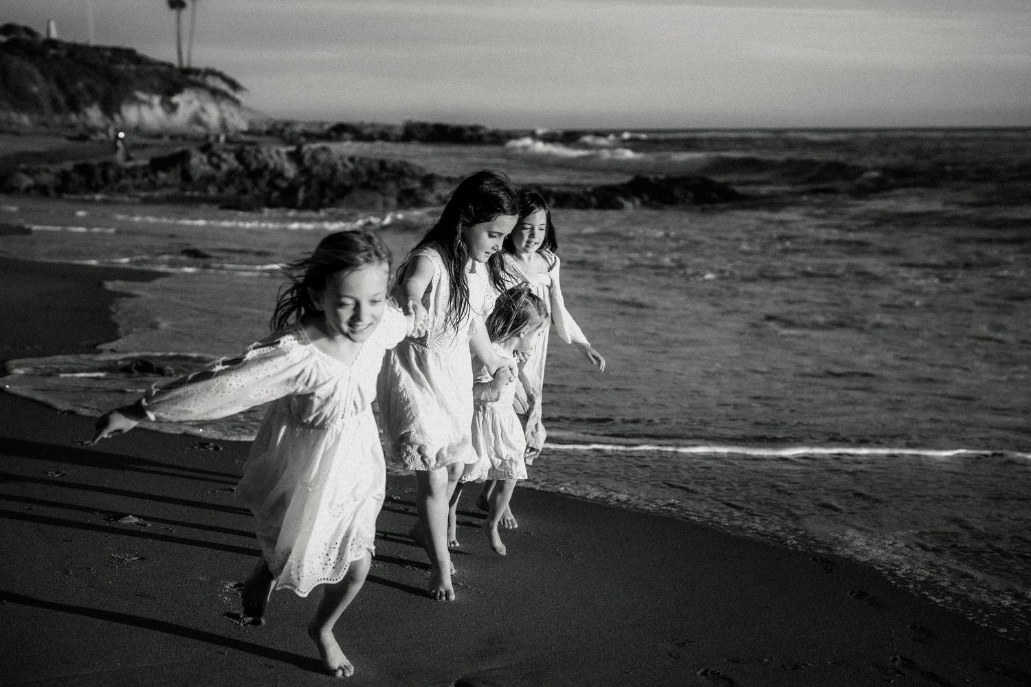 Black and white photo of four girls running joyfully along a beach shoreline.