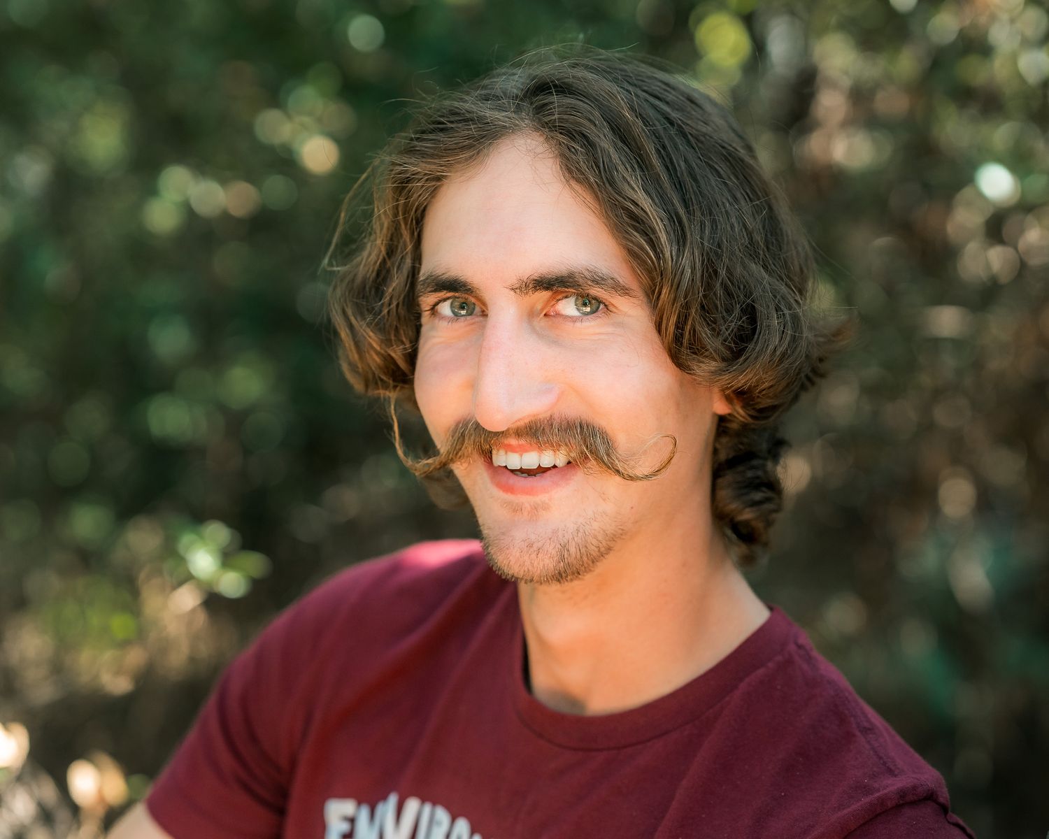 Young man with a mustache and wavy hair smiling in a maroon t-shirt with green foliage behind.