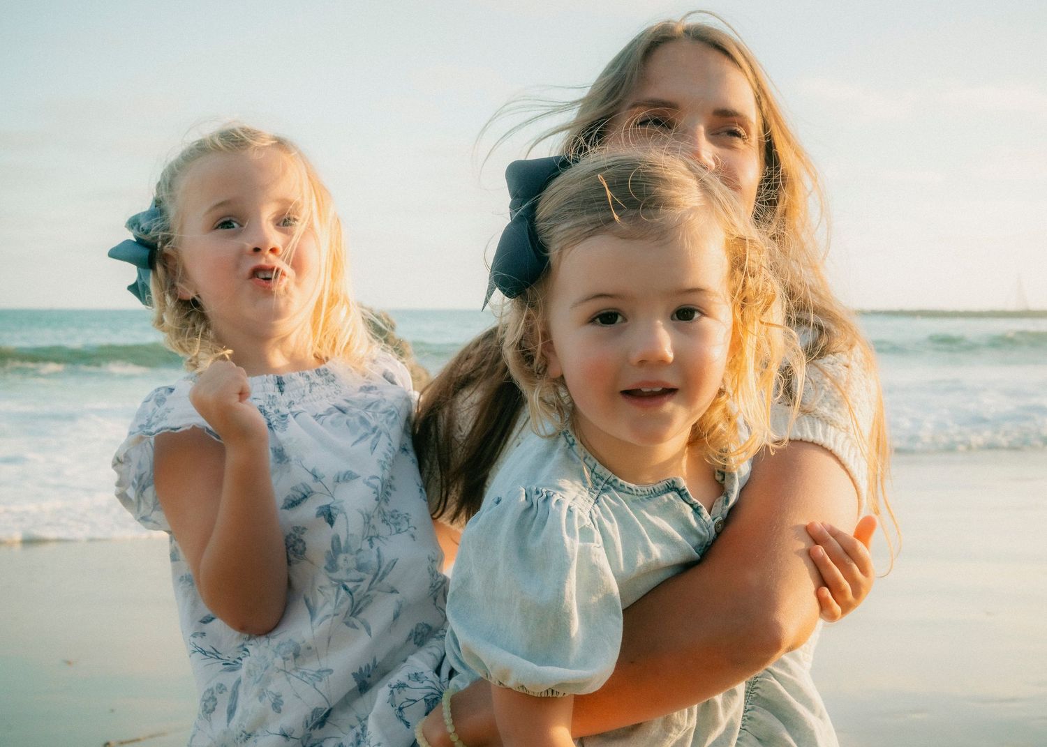 Two young blonde sisters with blue bows posing playfully together at the beach.