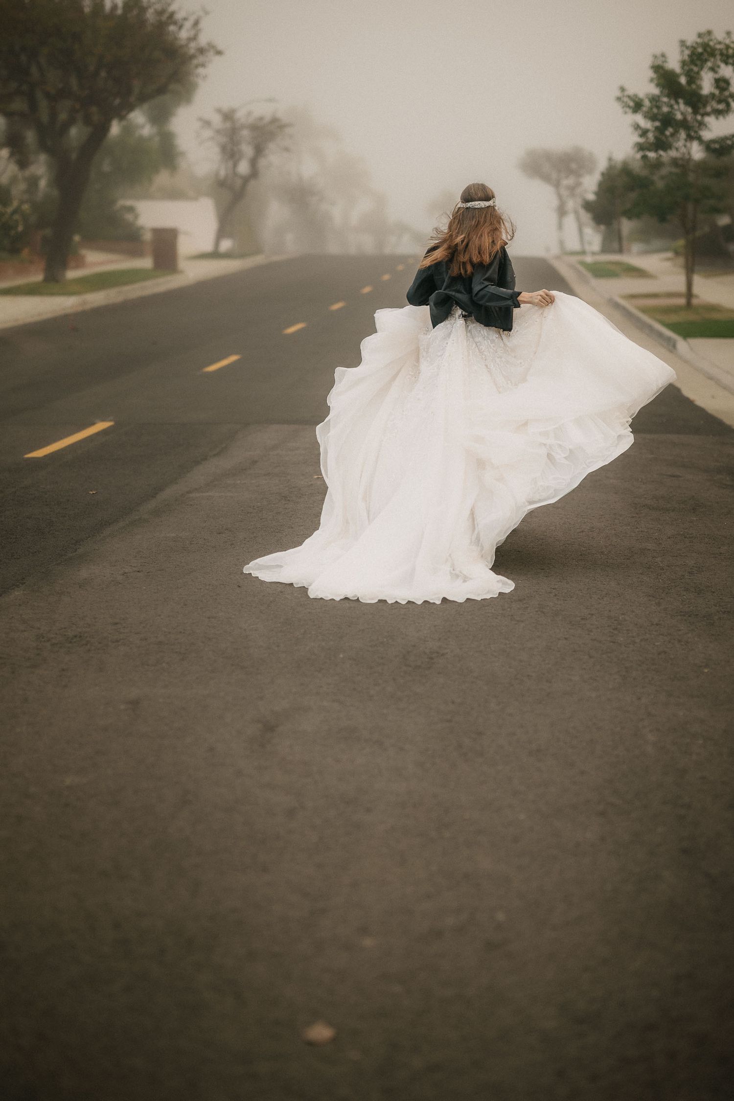 Bride in white gown and leather jacket twirling on foggy road with flowing train.