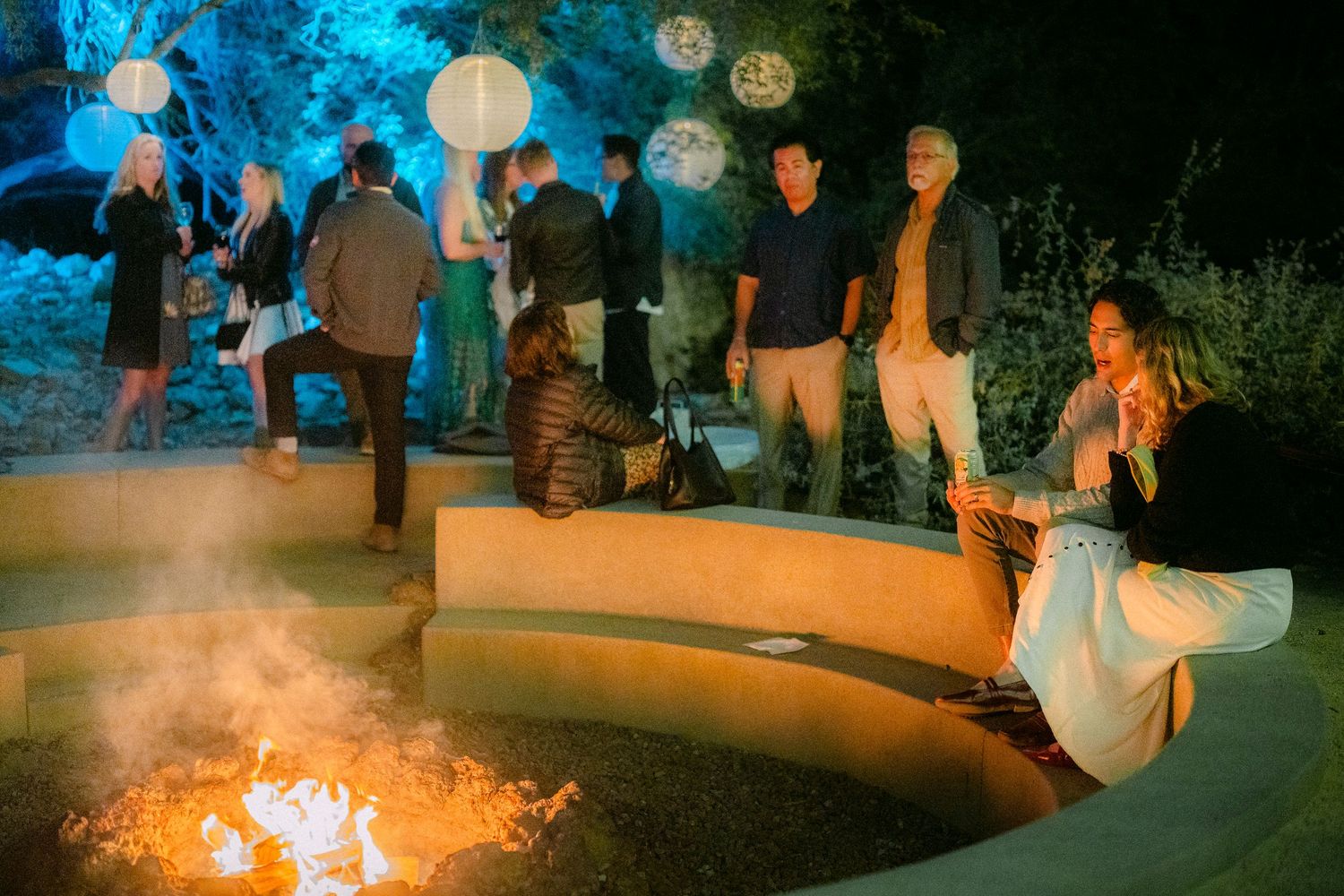 Guests socializing around a fire pit under lanterns at an elegant outdoor evening event.