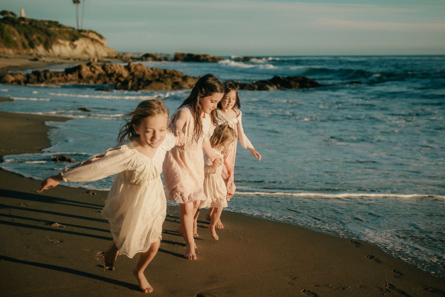 Four girls in white and pink dresses running playfully along a sunny beach shore.