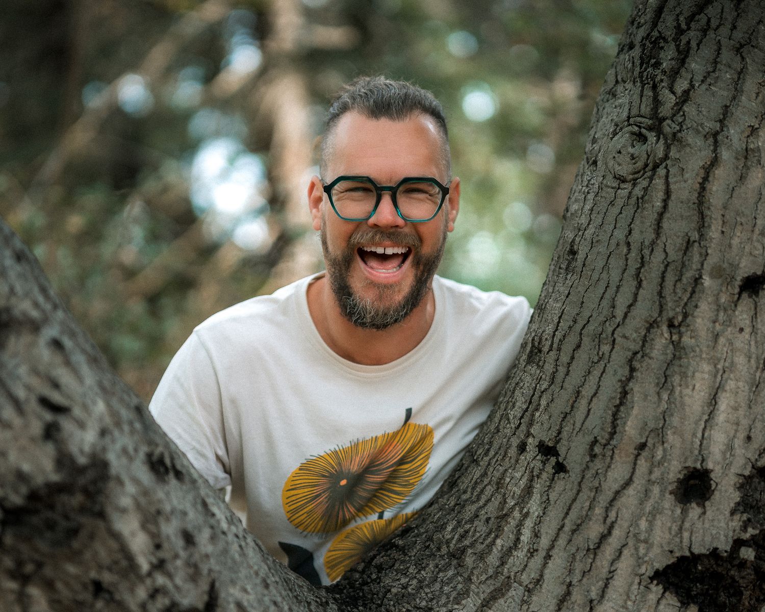 Bearded man with glasses laughing between tree trunks wearing a white mushroom graphic tee outdoors.