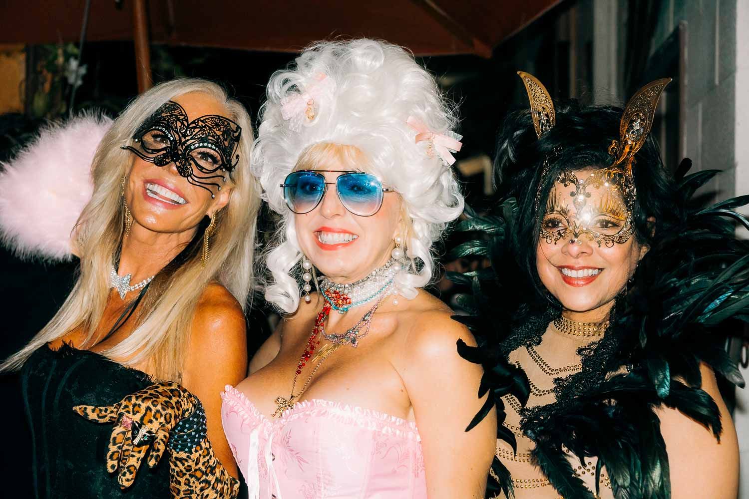 Three women in masquerade masks and costumes posing together at a festive party.
