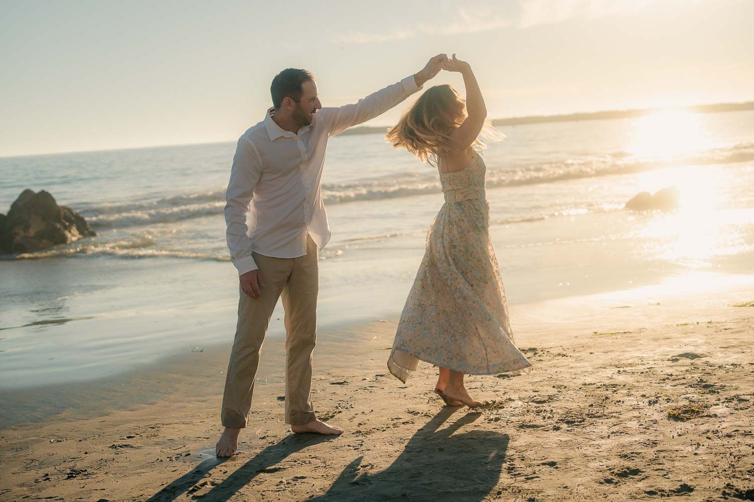 Couple dancing playfully on sandy beach at sunset, woman in lace dress being twirled.