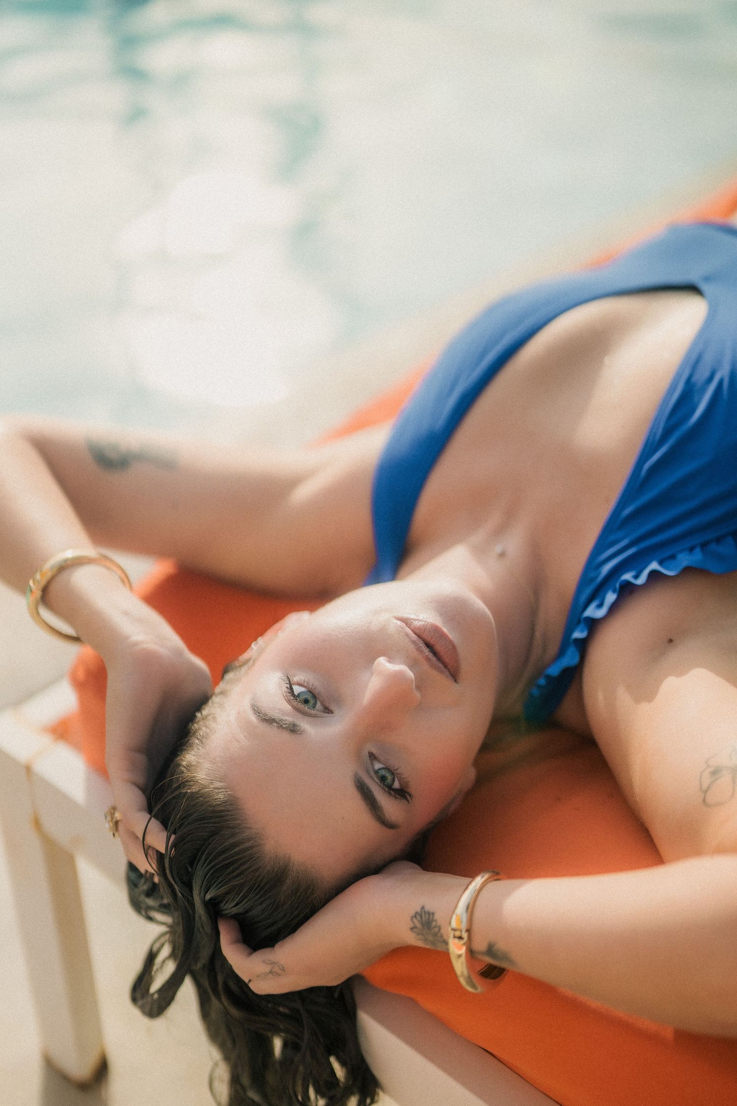 Woman in blue swimsuit relaxing on orange pool lounger with tattoos visible.