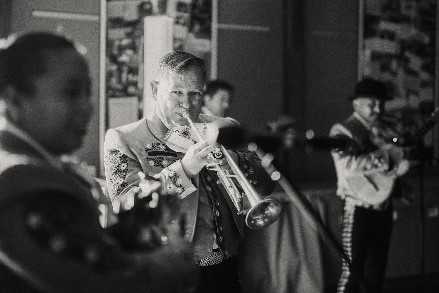 Black and white photo of musicians playing trumpets in mariachi-style uniforms at an indoor event.