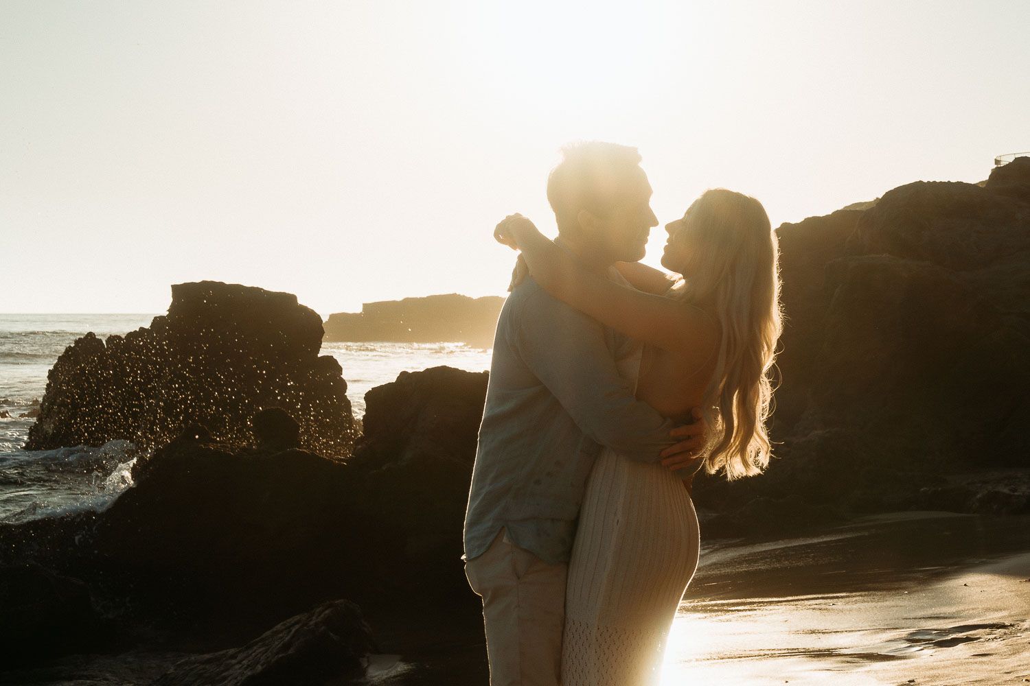 Couple embracing romantically on beach at sunset with rocky backdrop.