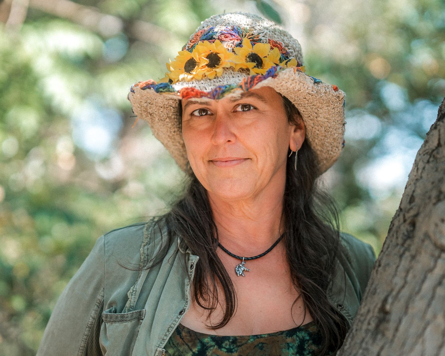 A woman in a floral straw hat and green jacket leans against a tree in a lush outdoor setting.