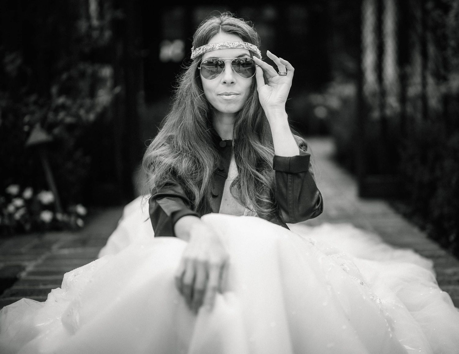 Black and white portrait of woman in tulle skirt and sunglasses sitting on garden pathway.