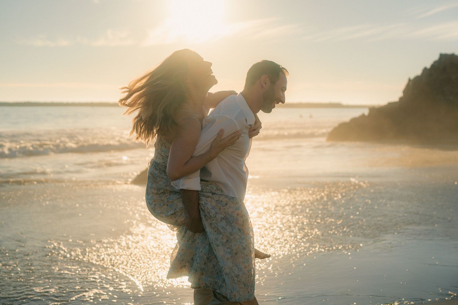 Man lifting woman joyfully on sparkling sunlit beach with golden hour ocean backdrop.