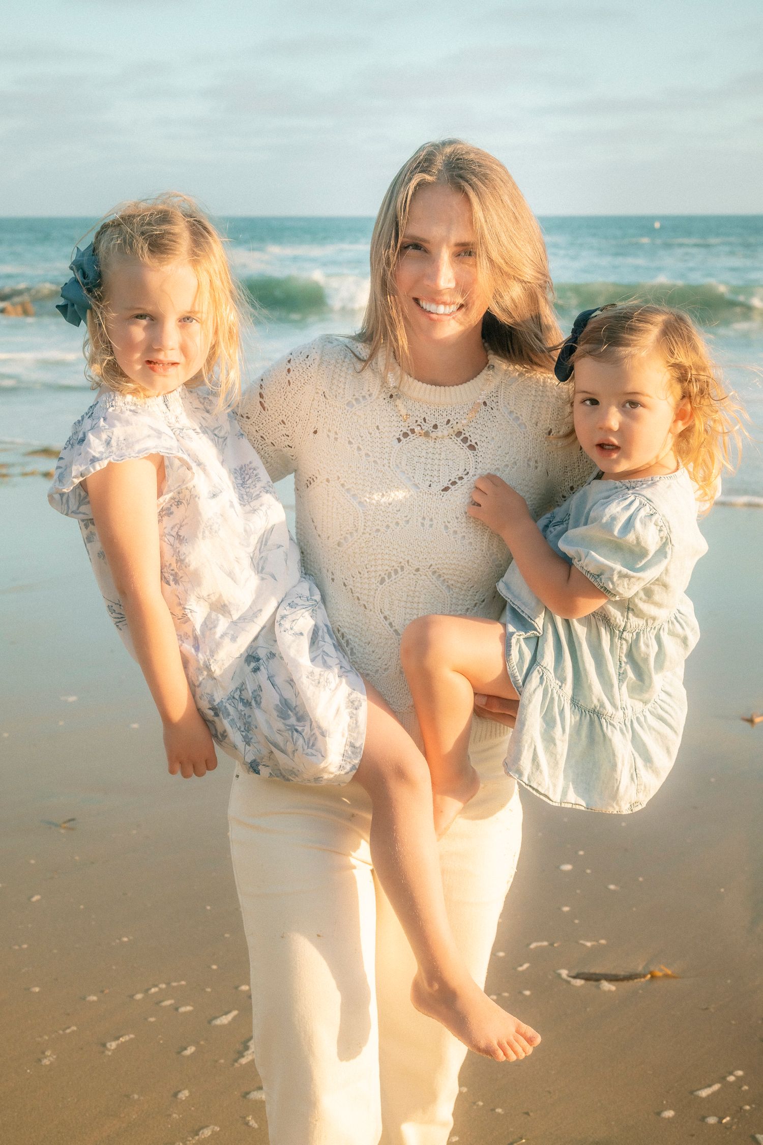 Blonde mother holding two young daughters on a sunny sandy beach by the ocean.