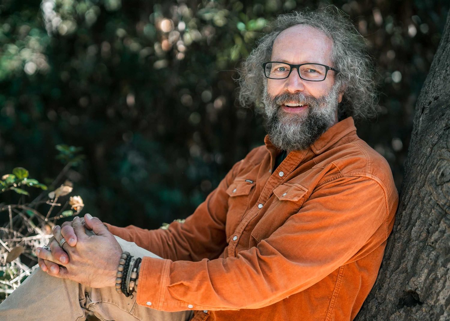 Smiling bearded man with curly hair and glasses in orange jacket leaning against tree outdoors.