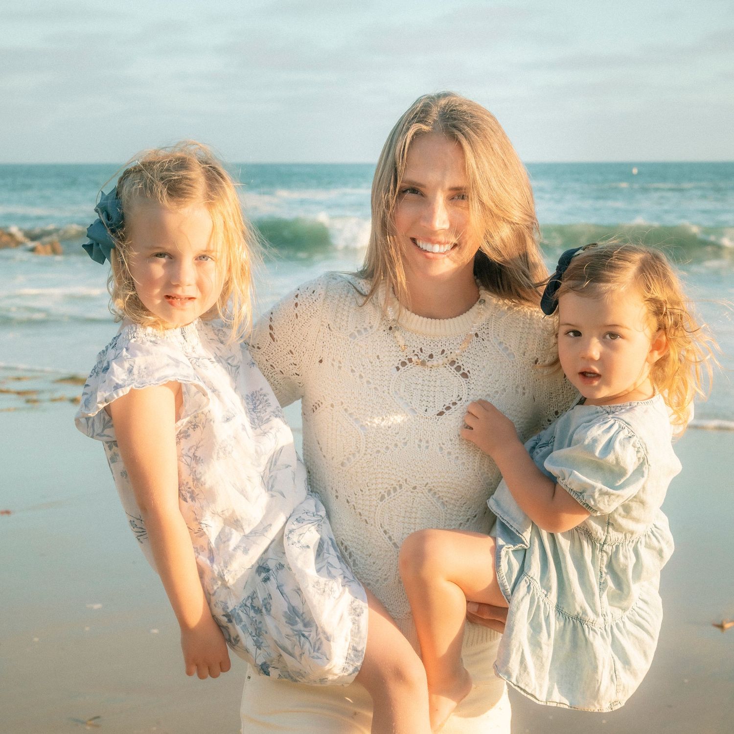 Smiling blonde woman holding two young girls on a sunny beach with ocean waves in the background.