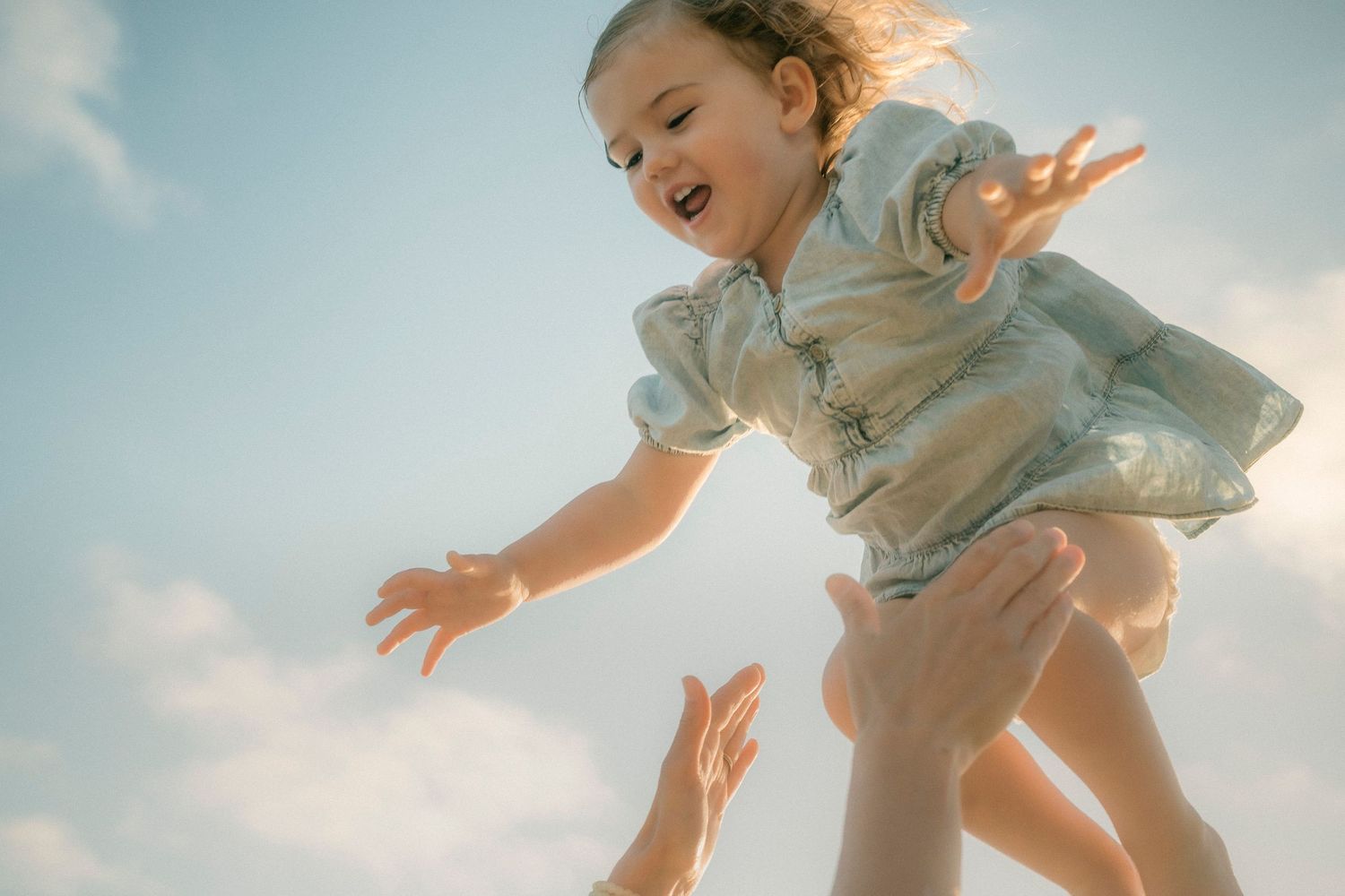Joyful toddler girl in floral dress being tossed in the air against blue sky.